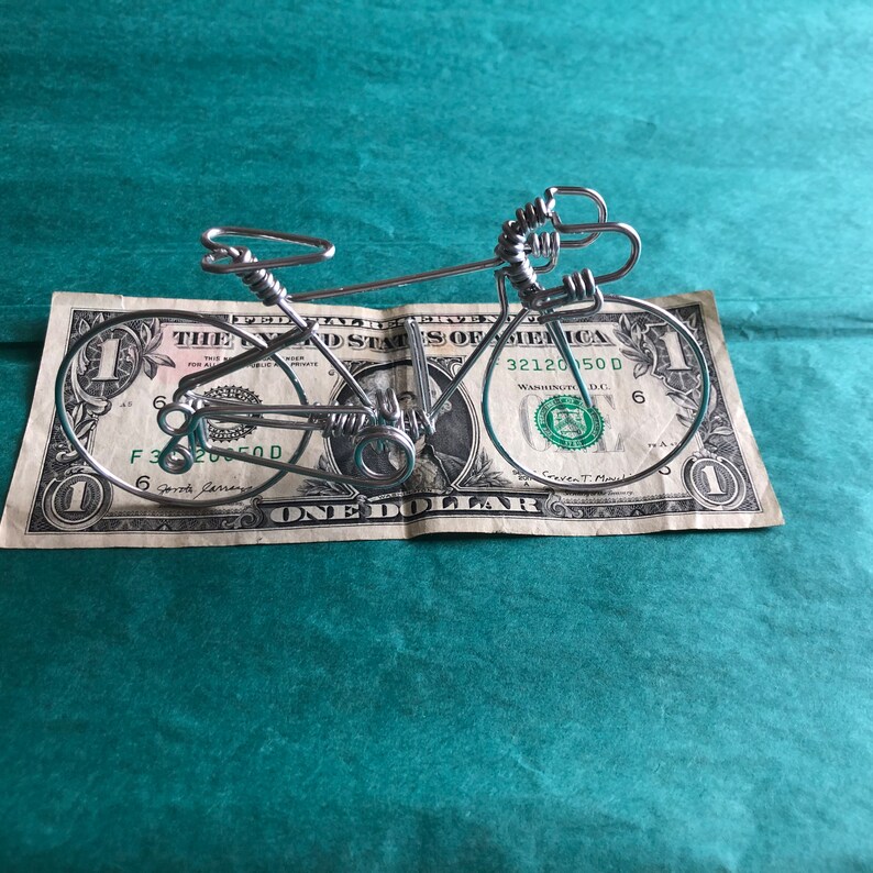 May include: A silver wire sculpture of a bicycle resting on a one dollar bill. The bill is folded in half and the bicycle is positioned on top of the bill. The bill features the image of George Washington.