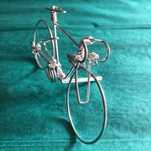 May include: A silver wire sculpture of a bicycle. The sculpture is made of thin wire that has been twisted and bent to create the shape of a bicycle. The bicycle has two wheels, handlebars, a seat, and pedals. The sculpture is on a green background.
