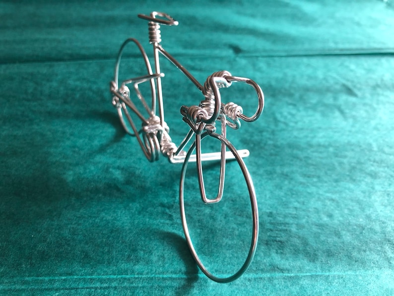 May include: A silver wire sculpture of a bicycle. The sculpture is made of thin wire that has been twisted and bent to create the shape of a bicycle. The bicycle has two wheels, handlebars, a seat, and pedals. The sculpture is on a green background.
