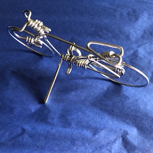May include: A silver wire sculpture of a bicycle. The sculpture is made from a single piece of wire that is bent and twisted to create the shape of a bicycle. The sculpture is on a blue background.