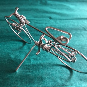May include: A silver wire sculpture of a bicycle. The sculpture is made of thin wire that has been twisted and bent to create the shape of a bicycle. The bicycle is on a stand and has two wheels.