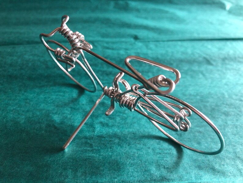 May include: A silver wire sculpture of a bicycle. The sculpture is made of thin wire that has been twisted and bent to create the shape of a bicycle. The bicycle is on a stand and has two wheels.