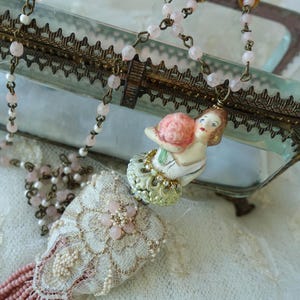 May include: A vintage necklace with a porcelain figurine pendant holding a pink flower. The necklace has pink beads and a decorative tassel. The jewellery is displayed near a glass and metal trinket box.