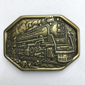 May include: A rectangular, antique brass-colored belt buckle featuring a detailed relief of a steam train. The train is depicted with intricate mechanical details, smoke, and the number "747" on top. The buckle has an octagonal border.