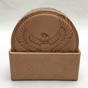 May include: A set of circular tan leather coasters featuring an embossed falcon with spread wings, housed in a matching rectangular tan leather holder with an embossed floral pattern on the front panel. The coasters are stacked vertically.