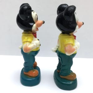Op de afbeelding: Twee vintage Mickey Mouse-beeldjes. Elke figuur heeft een zwart hoofd met grote oren, een geel shirt, een blauwe broek en oranje schoenen. De figuren staan op een tealkleurige basis en hebben een rode strik. De beeldjes staan naast elkaar.