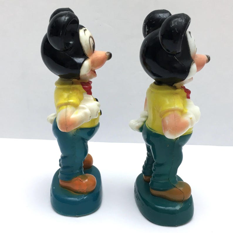 Op de afbeelding: Twee vintage Mickey Mouse-beeldjes. Elke figuur heeft een zwart hoofd met grote oren, een geel shirt, een blauwe broek en oranje schoenen. De figuren staan op een tealkleurige basis en hebben een rode strik. De beeldjes staan naast elkaar.