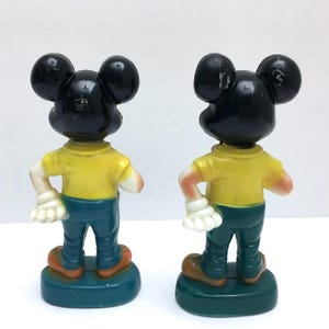 Op de afbeelding: Twee vintage Mickey Mouse beeldjes, van achteren bekeken. Elk figuur heeft een zwart hoofd, een geel shirt, een blauwe broek en bruine schoenen. Ze staan op een groene basis, met witte handschoenen en armen.