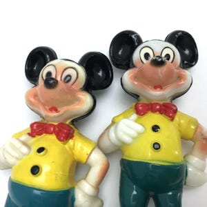 Op de afbeelding: Twee vintage Mickey Mouse beeldjes. Elke figuur heeft zwarte oren, een wit gezicht en een rode vlinderdas. Ze dragen gele shirts met zwarte knopen en teal broeken. De figuren zijn gemaakt van hard plastic.