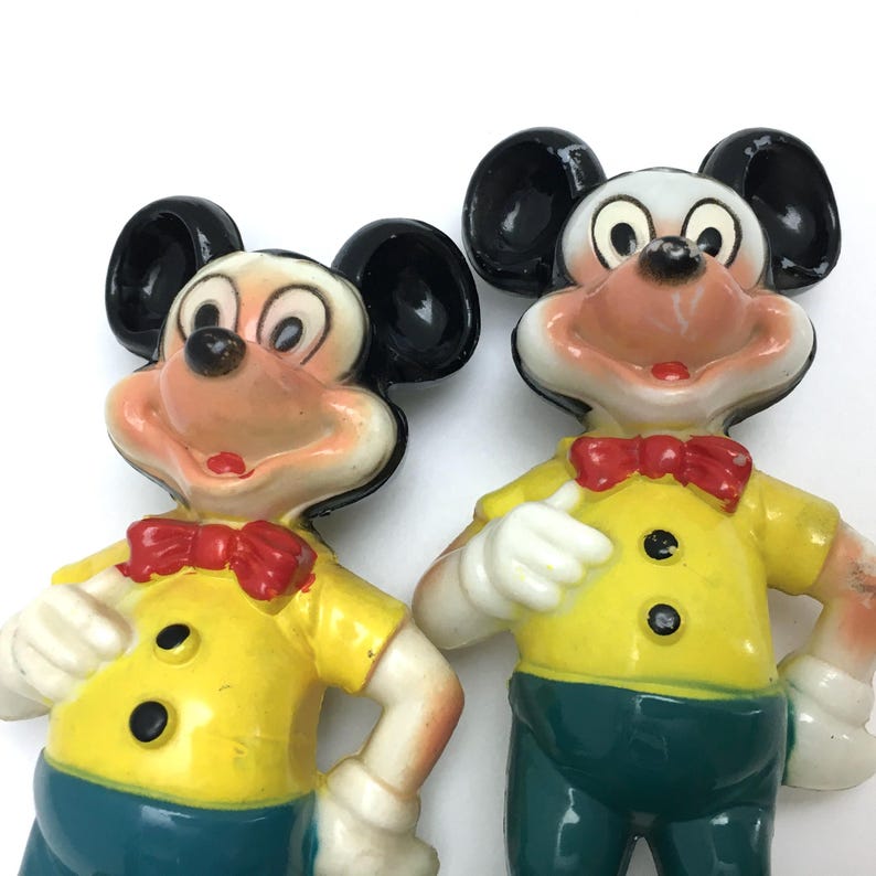 Op de afbeelding: Twee vintage Mickey Mouse beeldjes. Elke figuur heeft zwarte oren, een wit gezicht en een rode vlinderdas. Ze dragen gele shirts met zwarte knopen en teal broeken. De figuren zijn gemaakt van hard plastic.