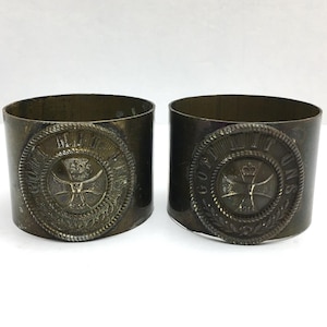 May include: Two antique brass napkin rings. Each ring features a circular emblem with a cross, crown, and text. The rings have a dark, aged patina, suggesting they are vintage or antique items. The design is detailed and ornate.