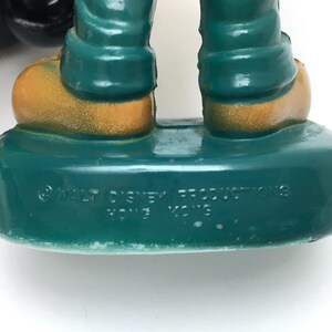 Op de afbeelding: Een close-up van een vintage beeldjevoet. De voet is zeegroen met de tekst "&copy; WALT DISNEY PRODUCTIONS HONG KONG" in het wit. De benen van het beeldje zijn zeegroen en de schoenen zijn goud.