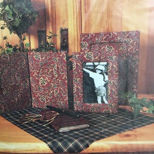 May include: A collection of decorative picture frames with a paisley pattern in red, green, and gold. One frame holds a black and white photo. A notebook, pen, and glasses rest on a plaid fabric.