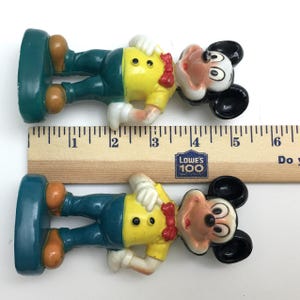 Op de afbeelding: Twee vintage Mickey Mouse-beeldjes. Elk beeldje is ongeveer 7,6 cm hoog. Mickey draagt een geel shirt, een blauwe broek en bruine schoenen. Een rode vlinderdas en witte handschoenen maken de look compleet. De beeldjes staan op een groene basis.