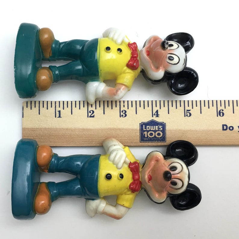 Op de afbeelding: Twee vintage Mickey Mouse-beeldjes. Elk beeldje is ongeveer 7,6 cm hoog. Mickey draagt een geel shirt, een blauwe broek en bruine schoenen. Een rode vlinderdas en witte handschoenen maken de look compleet. De beeldjes staan op een groene basis.