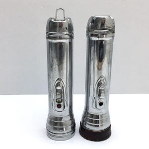 May include: Two vintage, cylindrical flashlights with a shiny, metallic finish. Each flashlight has a push-button switch, a handle loop, and a ribbed base. One has a red lens, the other a brown base.