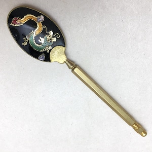 May include: A decorative spoon featuring a black enamel bowl with a gold, green, and purple dragon motif. The handle is made of a long, hexagonal brass-toned metal rod with a textured tip, connecting to the bowl with a smooth, tapered transition.