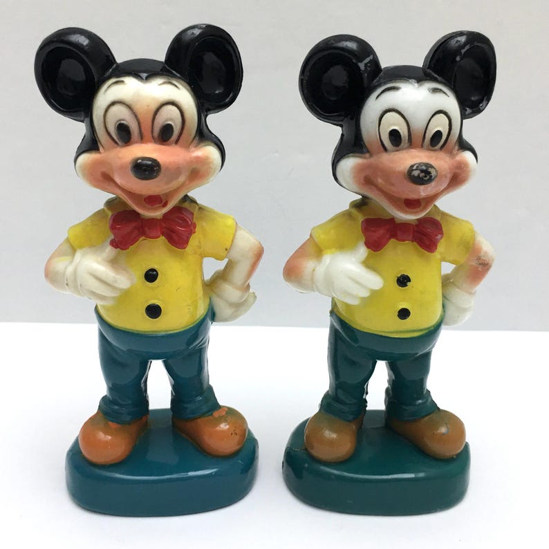 Op de afbeelding: Twee vintage Mickey Mouse beeldjes. Elk beeldje heeft zwarte oren, een wit gezicht en een rode vlinderdas. Ze dragen gele shirts, blauwe broeken en bruine schoenen. De beeldjes staan op een teal basis.