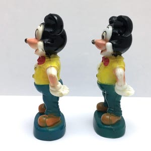 Op de afbeelding: Twee vintage Mickey Mouse beeldjes. Elke figuur heeft een zwart hoofd, een geel shirt, een blauwe broek en bruine schoenen. De figuren staan op een blauwe basis. De figuren staan naast elkaar.