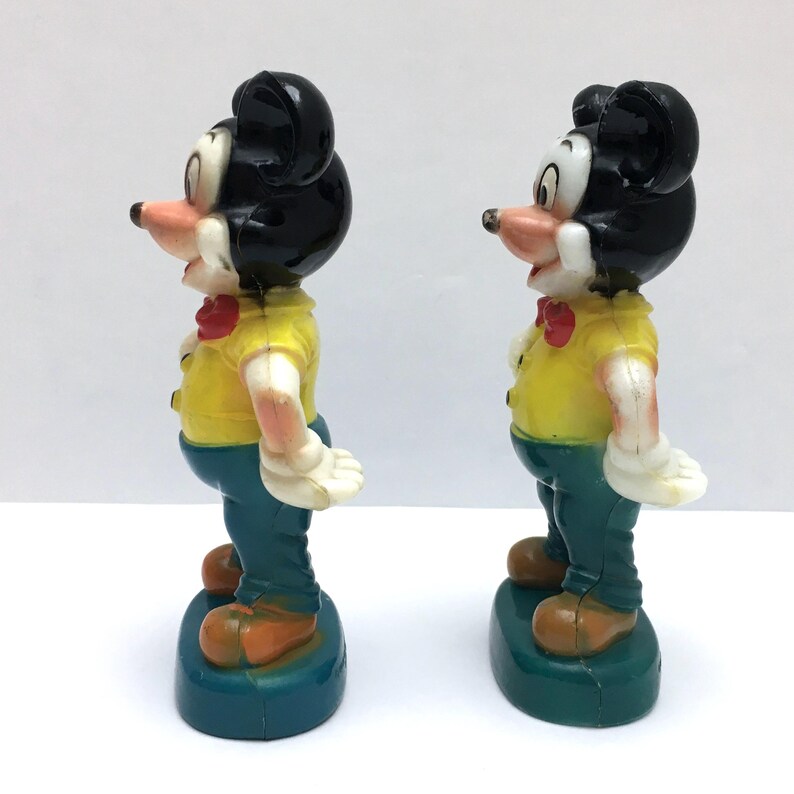 Op de afbeelding: Twee vintage Mickey Mouse beeldjes. Elke figuur heeft een zwart hoofd, een geel shirt, een blauwe broek en bruine schoenen. De figuren staan op een blauwe basis. De figuren staan naast elkaar.