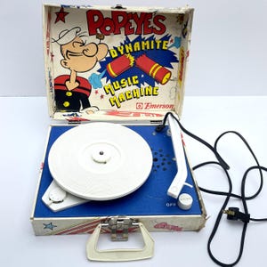 May include: Vintage Popeye's Dynamite Music Machine record player. The portable player has a white turntable, a blue base, and a white tone arm. The case features Popeye graphics and the Emerson logo. The case is white with red and blue accents.