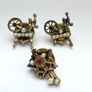 May include: Three vintage gold-toned brooches. Two brooches are shaped like spinning wheels with red center stones and faux pearls. The third brooch has a floral design with a red center stone and faux pearls. All three brooches are on a white background.