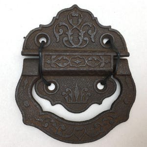 May include: A dark brown cast iron drop handle featuring an ornate, symmetrical relief pattern with scrolling details. The hardware consists of a rectangular mounting plate and a large, curved, decorative pull ring connected by two black wire loops.