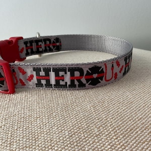May include: Gray dog collar with red and black accents. The collar has the words "HERO" and "U" printed on it, along with a red heart and a firefighter's axe.