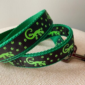 May include: Black and green dog leash with a repeating pattern of green lizards on a black background. The leash has a silver metal clip.