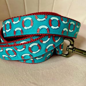 May include: A turquoise dog leash with a pattern of white and red life preservers. The leash has a red trim and a silver metal clasp. The leash is coiled, showing the pattern and the red underside.