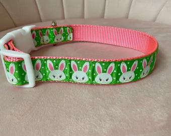 Puede incluir: Un collar rosa y verde para perros con una hebilla de plástico blanco. El collar presenta una cinta verde con lunares blancos y caras de conejitos de dibujos animados repetidas con orejas rosas. La parte interior del collar es rosa liso.