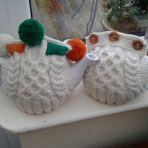 Puede incluir: Dos calienta-teteras de punto blanco con pompones y botones de diferentes colores. Un calienta-teteras tiene un pompón verde, naranja y blanco y el otro tiene un botón marrón.