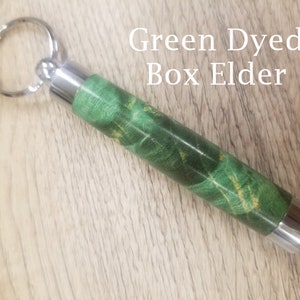 May include: A green-dyed Box Elder wood keychain with silver-colored metal accents. The wood grain features a mix of green and gold hues. The text "Green Dyed Box Elder" is visible.