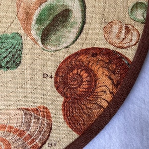 May include: A close-up of a beige fabric with a brown border. The fabric features a pattern of various seashells in shades of brown, green, and white. The text "D4" and "H5" is visible on the fabric.