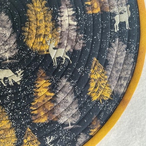 May include: A round, quilted table mat with a black background and a gold and white pattern of trees and deer. The mat has a gold border.