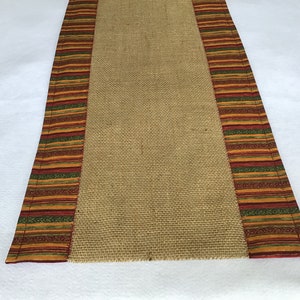 May include: A brown burlap table runner with a colorful striped border. The border features red, orange, green, and yellow stripes with a repeating pattern.