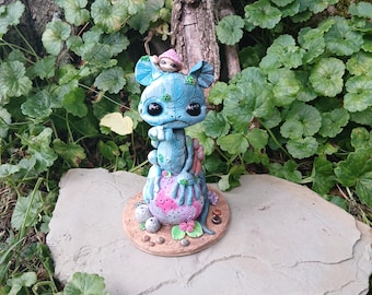 Op de afbeelding: Een grillig blauw beeldje met grote zwarte ogen, een kleine hoed en een roze hartvormig ontwerp. Het beeldje staat op een stenen plaat omringd door groen, waardoor een fantasietentoonstelling ontstaat.