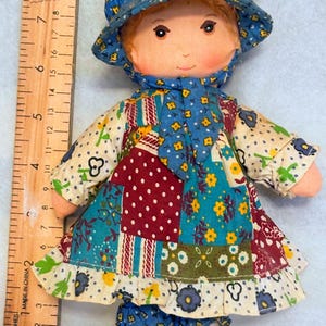 May include: A fabric doll wearing a blue bonnet with yellow flowers and a patchwork dress featuring red, white, and blue floral patterns. A wooden ruler placed beside the doll indicates a height of approximately 8.5 inches from head to toe.