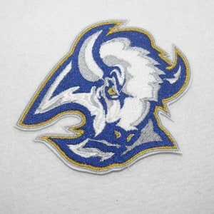 May include: A blue and gold embroidered patch featuring a white buffalo head with a fierce expression. The buffalo is surrounded by a blue and gold design.
