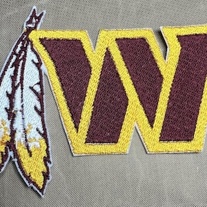 May include: An embroidered patch with a burgundy "W" outlined in yellow. A white and yellow feather design is on the left side of the patch.
