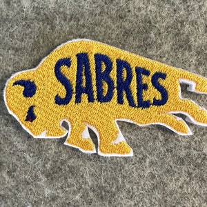 May include: Yellow embroidered patch shaped like a buffalo with the word "SABRES" in blue embroidery.