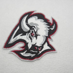 May include: Embroidered patch featuring a stylized buffalo head. The design uses black, white, gray, and red colors. The patch has a red border and is likely for apparel or accessories.