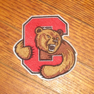 May include: A red embroidered patch with a white outline featuring a brown bear with a white muzzle and teeth. The bear is holding a red letter "C" in its paws.