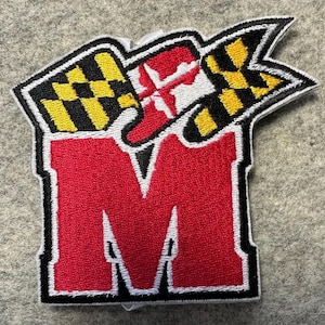 May include: Embroidered patch featuring a large red "M" with a white outline. Above the letter are two flags with a black and yellow checkered pattern and a red and white cross design. The patch has a black border and is set against a gray background.