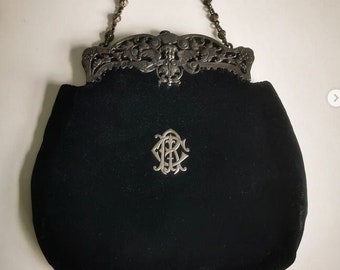 Könnte beinhalten: Eine schwarze Samt-Abendtasche mit einer silbernen Kettenkette und einem verzierten silbernen Verschluss. Die Tasche hat ein silbernes Monogramm.