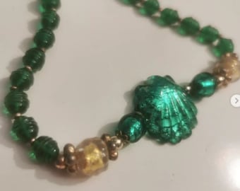 Könnte beinhalten: Eine Halskette aus grünen Glasperlen mit einem goldenen und grünen Muschel-Anhänger. Die Halskette besteht aus kleinen, runden Perlen und hat einen zarten, femininen Look.