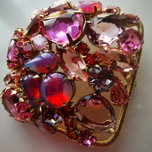 May include: A large, gold-toned brooch featuring a cluster of pink, red, and purple rhinestones. The brooch is square-shaped and has a textured metal frame.