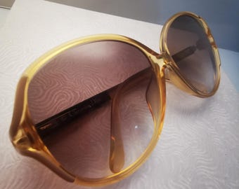 Könnte beinhalten: Eine Sonnenbrille in Goldtönen mit braunen Gläsern. Die Sonnenbrille hat einen großen, abgerundeten Rahmen und der Markenname "Christian Dior" ist auf dem linken Glas sichtbar.