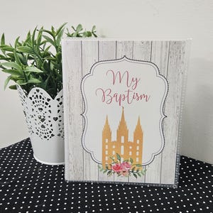 May include: A clear photo album with a cover featuring the text "My Baptism" in cursive script. The cover has a wood-grain background and a gold illustration of a building with three spires, accented with pink flowers.