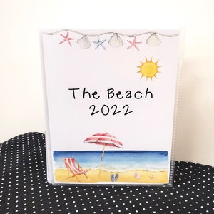May include: White photo album with a beach theme. The cover features a sun, beach umbrella, beach chair, and seashells. The text "The Beach 2022" is printed on the cover.